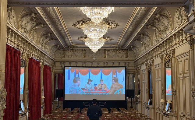 Écran de cinéma centré dans un salon aux décors dorés et aux lustres.